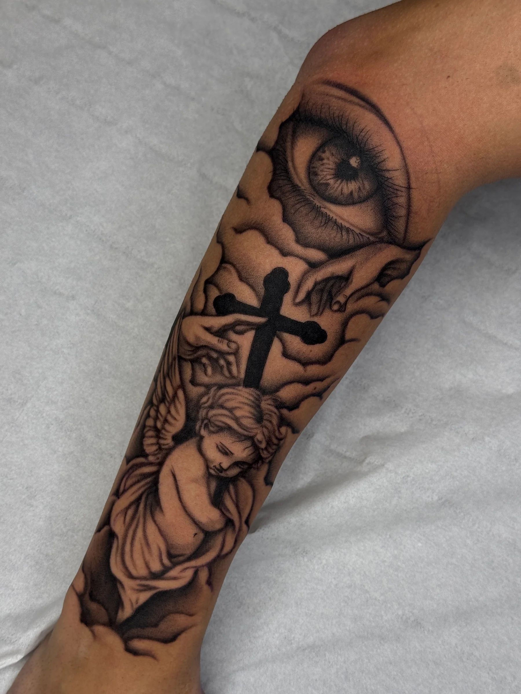 Un ojo entre nubes, una cruz y un angelito en el antebrazo